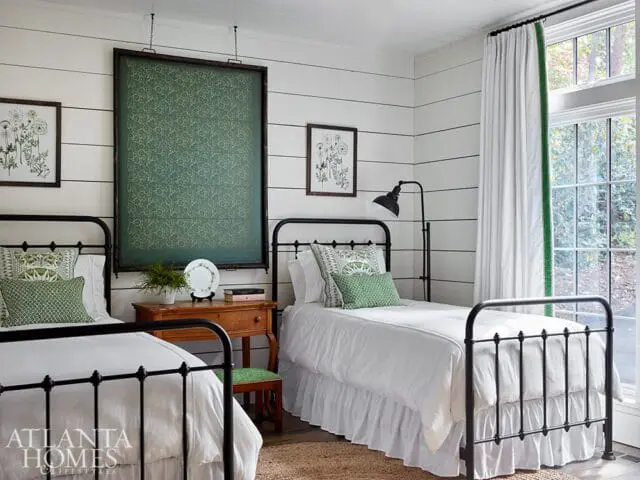 Bedroom with two twin beds featuring black metal frames, white bedding, and green patterned pillows. White shiplap walls are adorned with botanical prints and a large framed green textile.