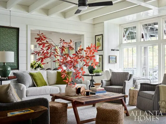 Living room with white shiplap walls, coffered ceiling, and large windows. Features a white sofa with green and patterned pillows, a rustic wood coffee table, and striped armchairs.