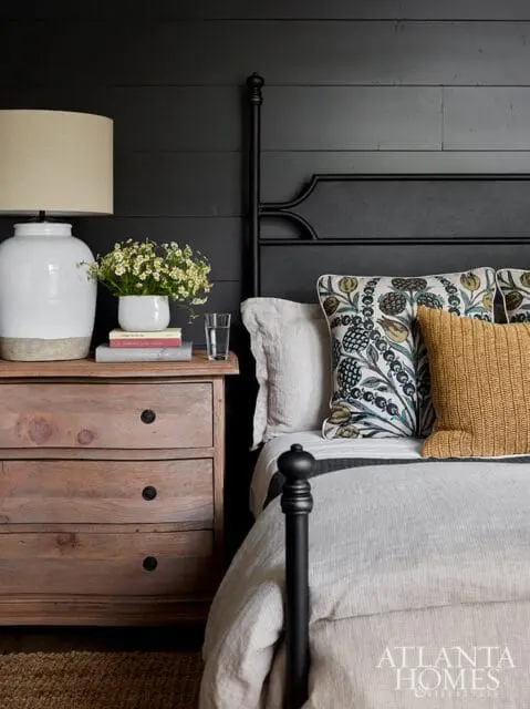 Bedroom with black shiplap walls, a dark metal bed frame, and a wooden nightstand with a large white lamp and a vase of white flowers.