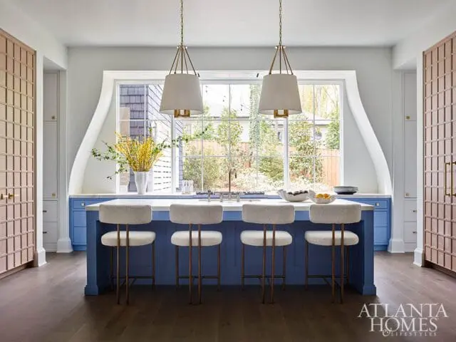 Blue kitchen island with white marble countertop and four upholstered stools, flanked by tall cabinets with grid detail, under a large window with pendant lights.