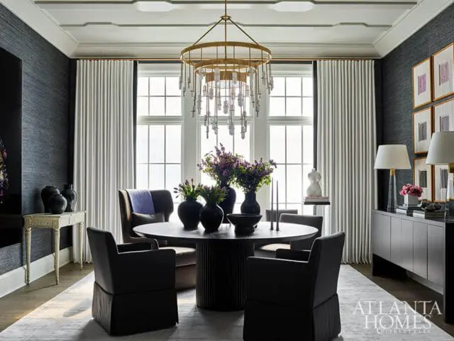 Dark textured wallpaper dining room with round fluted pedestal table, upholstered chairs, large windows with white drapes, and a gold chandelier.