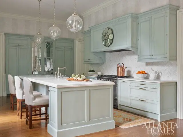 Light blue kitchen with white marble countertops, a large island with seating, glass globe pendant lights, and a copper kettle on the stove.