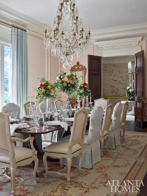 Formal dining room with a large wooden table set for a meal, upholstered chairs, a crystal chandelier, and floral arrangements.