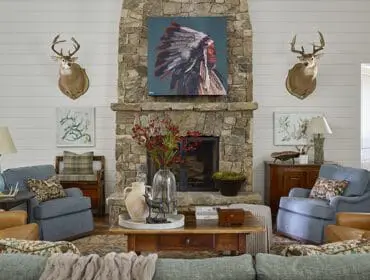 Living room with stone fireplace, mounted deer heads, and leather and upholstered armchairs. A painting of a Native American chief hangs above the fireplace.