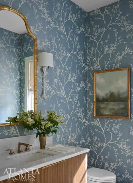 Powder room with blue floral wallpaper, marble vanity with brass fixtures, gold mirror, and a landscape painting.