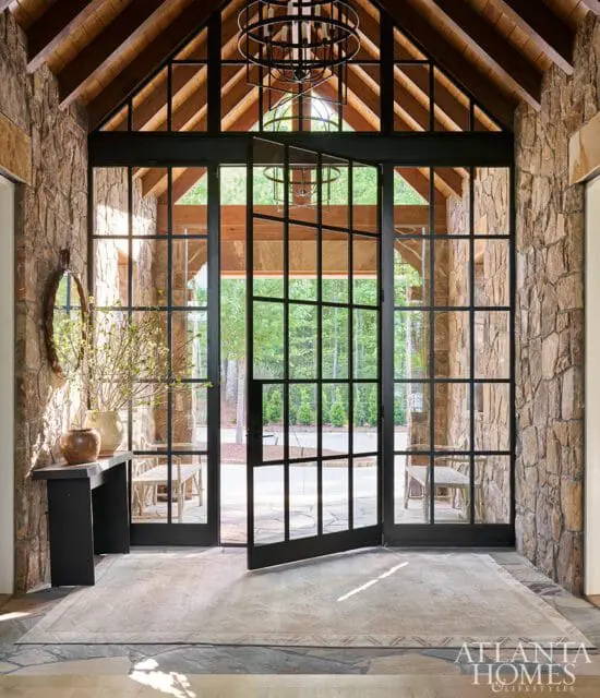 Entryway with stone walls, wood-beamed vaulted ceiling, and large black-framed glass doors opening to a patio. A circular chandelier hangs overhead.