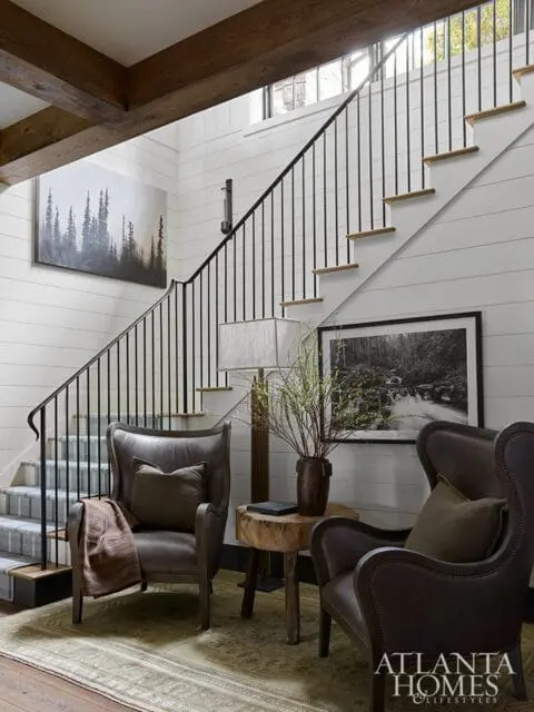 Interior staircase with shiplap walls, dark wood beams, black metal railing, and two leather wingback chairs with throw pillows and blankets.