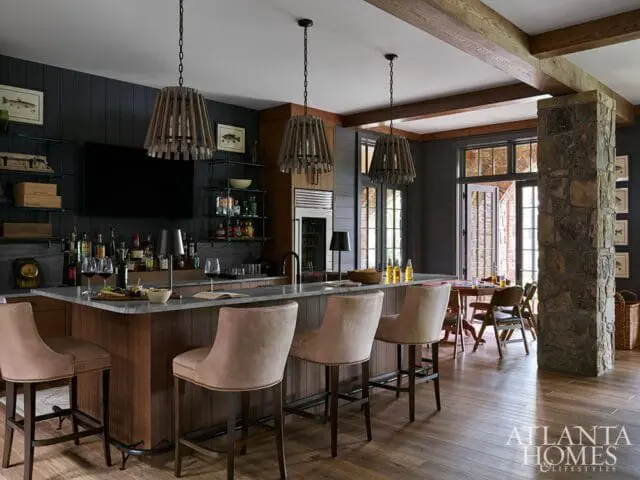 Bar area with gray stone countertop, dark wood cabinetry, and upholstered bar stools. Wooden pendant lights hang above. A stone pillar and dining area are visible in the background.