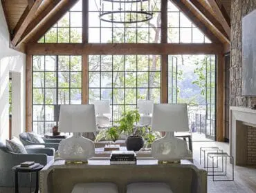 Living room with vaulted wood beam ceiling, large windows overlooking trees, stone fireplace, and light blue upholstered chairs.