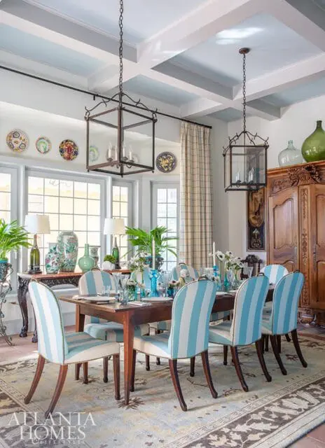 Dining room with a wooden table set for a meal, surrounded by chairs upholstered in blue and white stripes. A large wooden armoire stands against the wall, and two lantern-style chandeliers hang from the coffered ceiling.