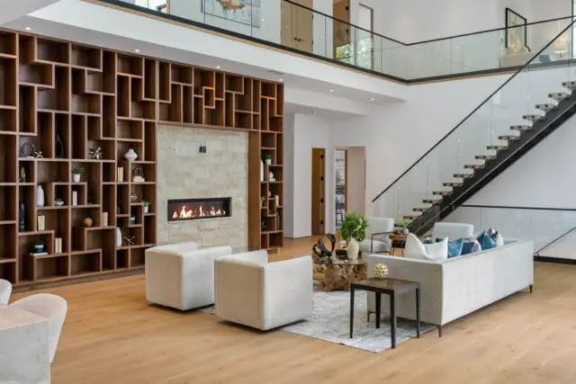 Modern living room with a large geometric wood bookshelf, stone fireplace with a fire, light gray sectional sofa with blue and white pillows, and a floating staircase with glass railings.