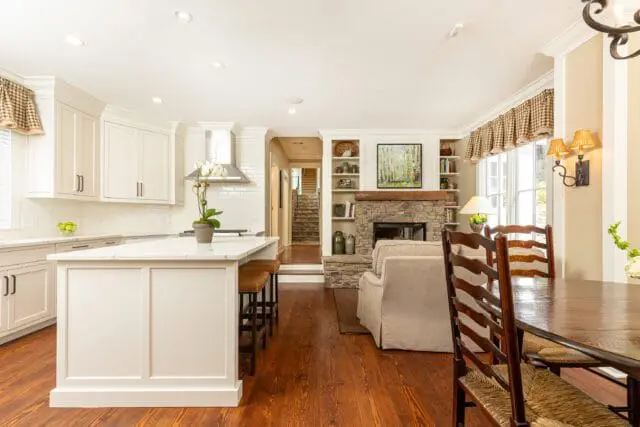 Open-concept kitchen and living area with white cabinetry, marble island, stone fireplace, and dining table with ladder-back chairs.