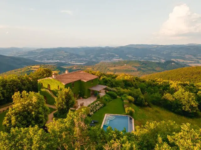 Vine-covered house with a terracotta roof sits on a hillside overlooking a valley, with a swimming pool in the foreground and lush green trees surrounding the property.