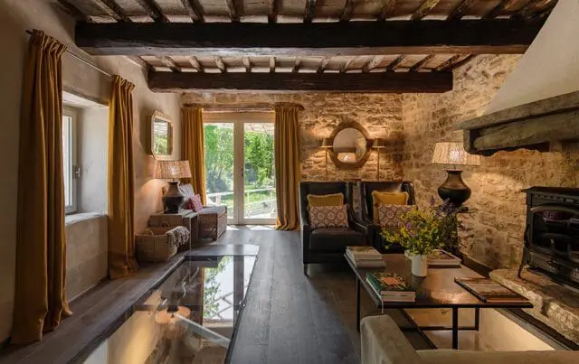 Living room with stone walls, exposed wooden beams, dark wood floors, mustard yellow curtains, two leather armchairs with patterned pillows, and a fireplace.