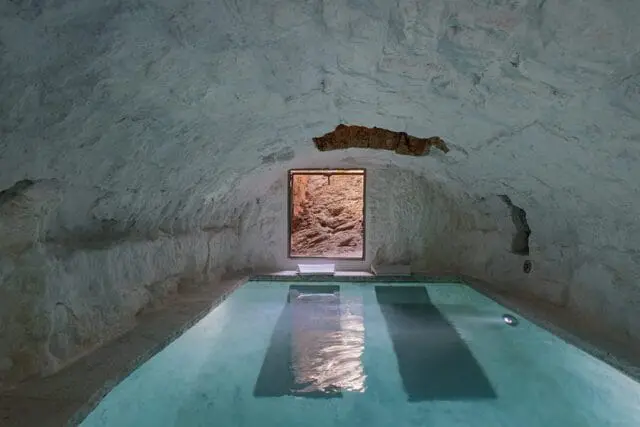 Indoor pool with rough, white-washed cave-like walls and ceiling, and a rectangular window looking out to a rocky exterior.