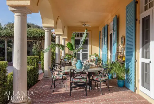 Covered patio with columns, yellow stucco walls, and bright blue shutters. A round stone table with wrought iron chairs is set with floral cushions.