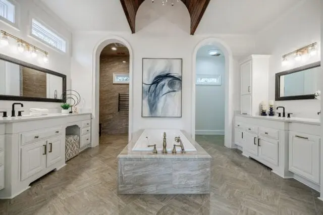 White bathroom with a freestanding soaking tub, dual vanities with white countertops and black faucets, and arched doorways leading to a tiled shower and a separate room.