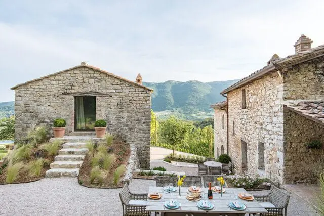 Stone buildings with tile roofs and a dining table set for a meal on a gravel patio, overlooking a lush green valley and mountains.