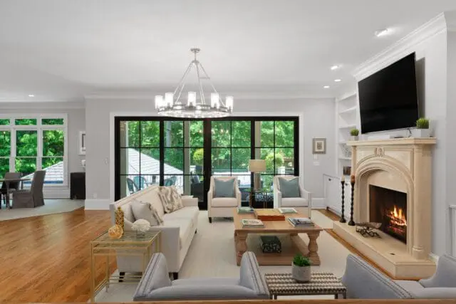 Living room with a light gray rug, cream sofa, two cream armchairs, and a light gray sofa. A large stone fireplace with a fire burning is on the right. Large windows overlook a lush green outdoor space.