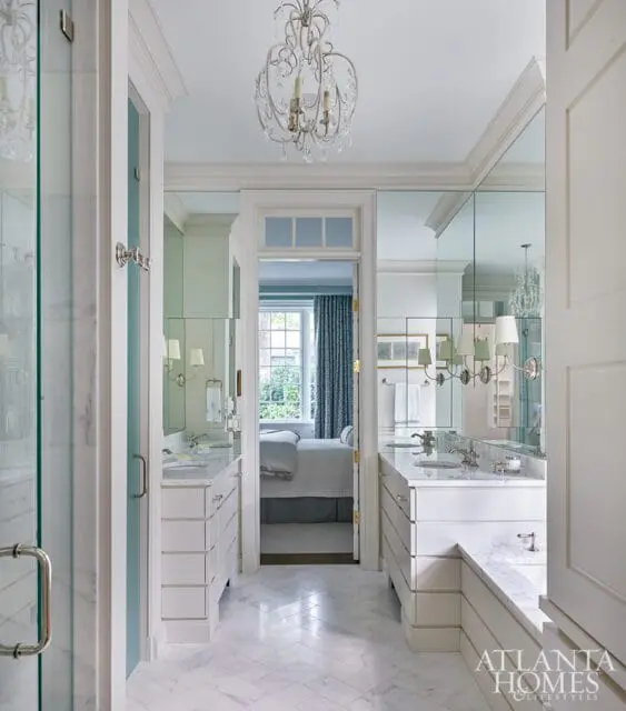 White marble bathroom with dual vanities, herringbone marble floors, and a view into a bedroom with blue patterned curtains.