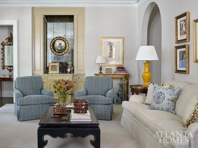 Living room with two blue patterned armchairs, a cream sofa with patterned pillows, a black coffee table, and a yellow lamp on a gold console table.