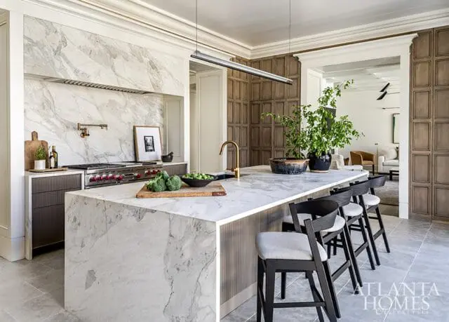 Modern kitchen with a large marble island, marble backsplash, stainless steel range, and dark wood paneled walls.