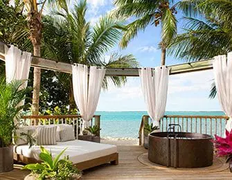 Wooden balcony with white curtains, daybed, and copper tub overlooking the ocean and palm trees.