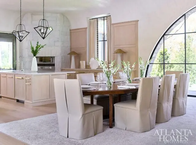 Dining room with a wooden table set for a meal, surrounded by upholstered chairs. A kitchen island with a stone fireplace is visible in the background, along with a large arched window.