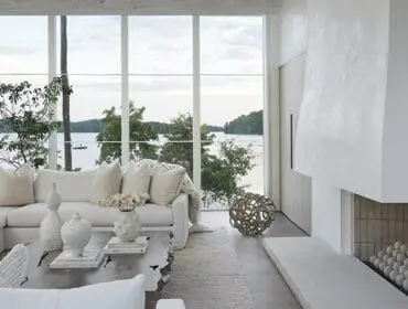 Living room with white sectional sofa, textured pillows, and a large abstract coffee table, overlooking a lake through floor-to-ceiling windows.