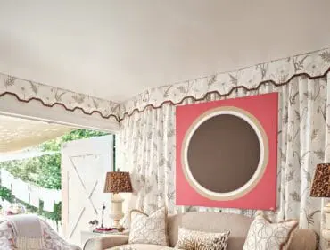 Living room with a neutral sofa, patterned armchairs, a glass-top coffee table, and abstract art on the wall. Doors open to an outdoor space.