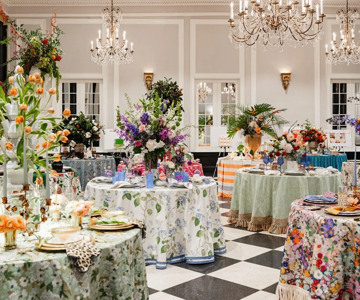 Elegant room with multiple round tables set for a formal event, featuring floral tablecloths, elaborate floral arrangements, and ornate chandeliers.