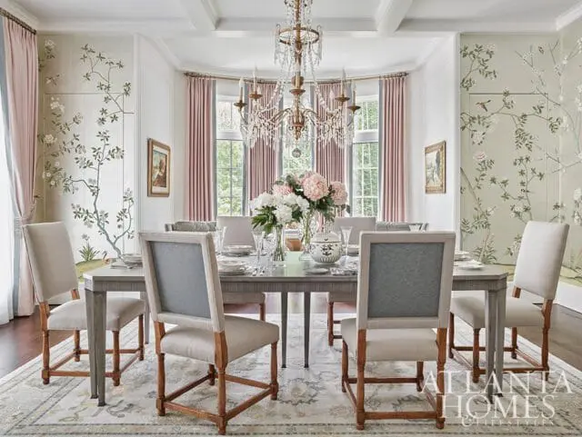 Dining room with a grey rectangular table set for a meal, surrounded by upholstered chairs. The walls feature a scenic mural with birds and flowering branches, complemented by pink drapes and a crystal chandelier.