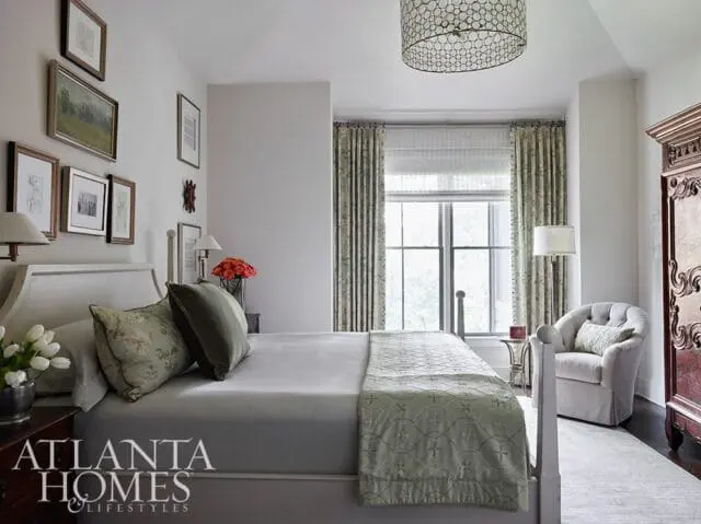 Bedroom with a white bed, floral curtains, a tufted armchair, and a large ornate armoire. Artwork hangs on the wall above the bed.