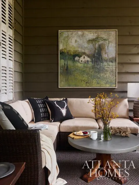 Covered porch with sectional sofa, concrete coffee table, and landscape painting. Pillows feature a deer head and geometric pattern.