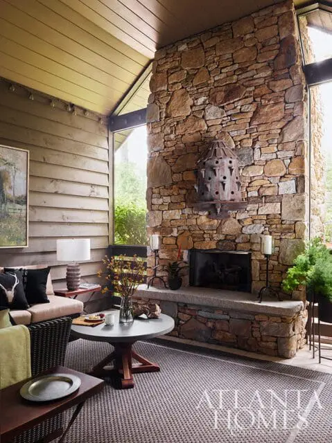 Covered porch with stone fireplace, wood paneled walls, and wicker furniture. A round coffee table sits on a patterned rug.