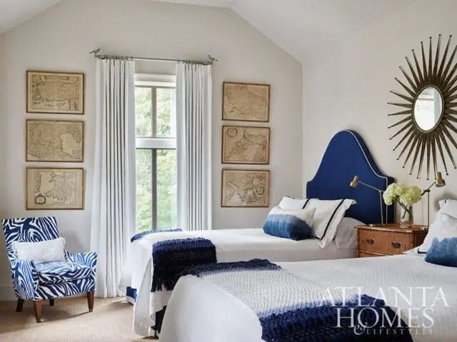 Bedroom with two twin beds featuring navy blue headboards, white bedding, and navy and white throws. Walls are adorned with framed antique maps. A blue and white zebra-print armchair sits in the corner.