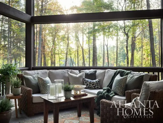 Screened porch with wicker sectional sofa, patterned pillows, and a wooden coffee table overlooking a sunlit forest