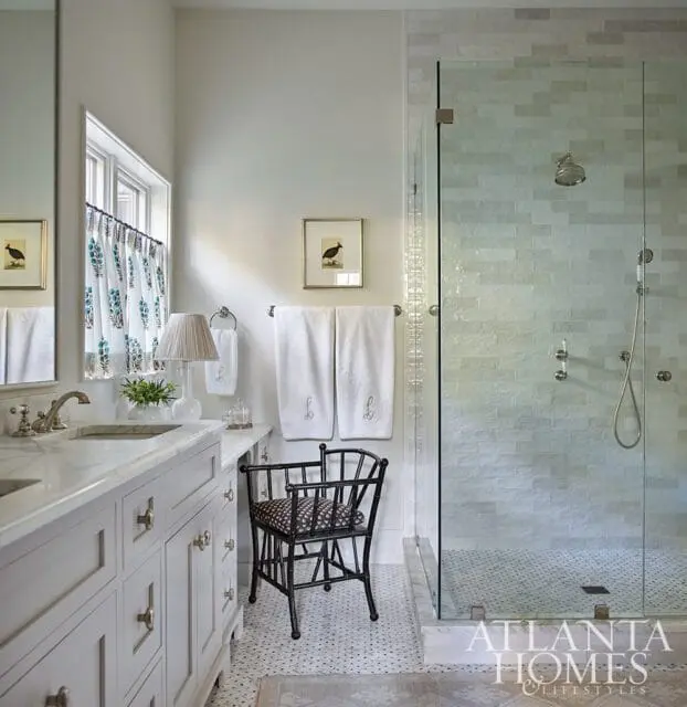 Light gray double vanity with marble countertop and brass hardware, glass-enclosed shower with white subway tile, and black accent chair with patterned cushion.