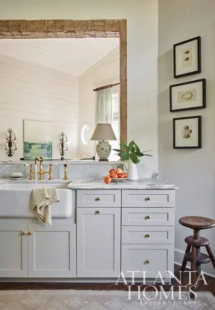 Light gray kitchen cabinets with brass hardware, white farmhouse sink, marble countertops, and framed botanical prints on the wall.