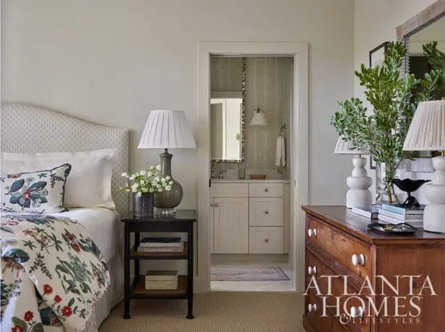 Bedroom with a patterned headboard and floral bedding, a dark nightstand with a lamp and flowers, and a view into a small bathroom with a vanity and patterned wallpaper. A wooden dresser with lamps and greenery is on the right.
