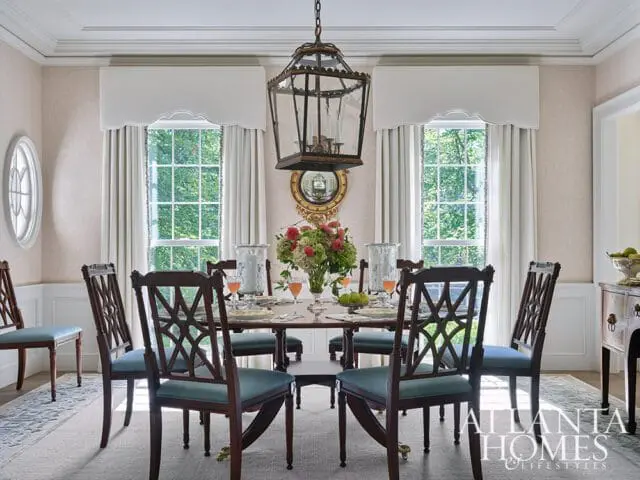 Dining room with a dark wood table set for a meal, surrounded by upholstered chairs with decorative backs. A large lantern hangs overhead, and windows with white drapes look out onto greenery.