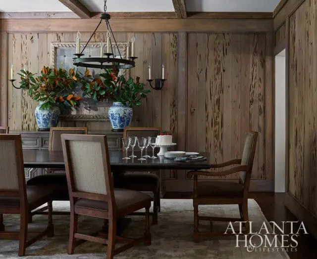 Dining room with dark wood paneled walls, a dark wood dining table set with wine glasses and a cake, upholstered chairs, and two large blue and white vases filled with greenery.