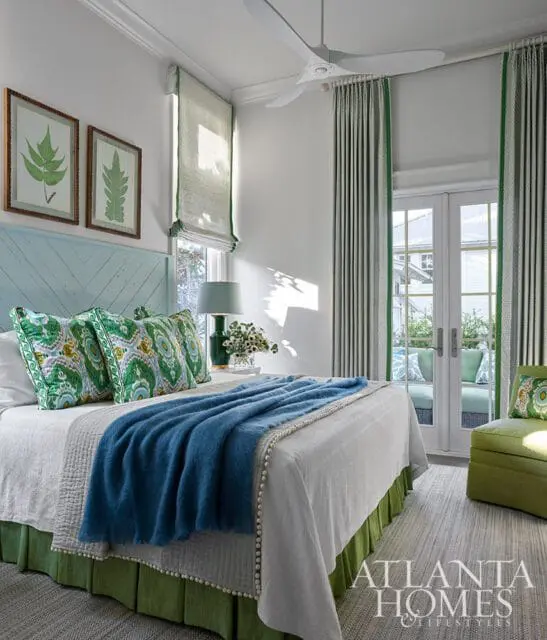 Bedroom with a light blue paneled headboard, green and white patterned pillows, a blue throw blanket, and green bed skirt. White walls, green and white striped curtains, and French doors leading to an outdoor patio.