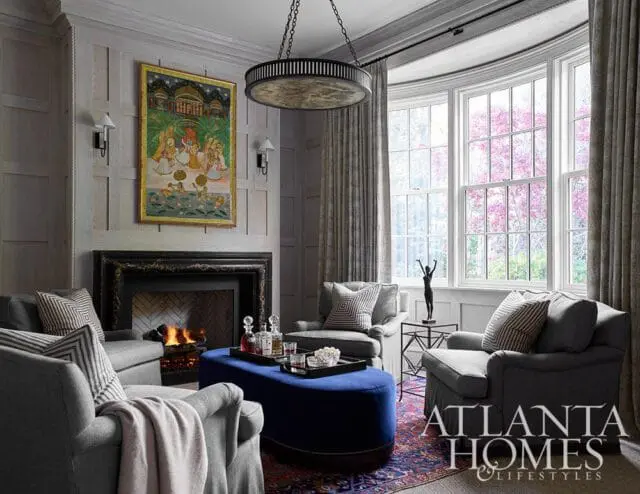 Living room with gray upholstered chairs, a fireplace with a black marble surround, a blue ottoman, and a painting above the fireplace. Large bay windows overlook a blooming tree.