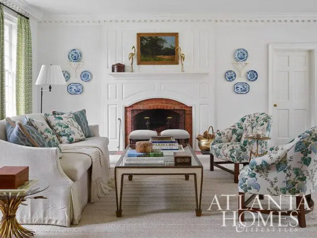 Living room with white fireplace, blue and white decorative plates, a sofa and two armchairs upholstered in floral fabric, and a glass-top coffee table.