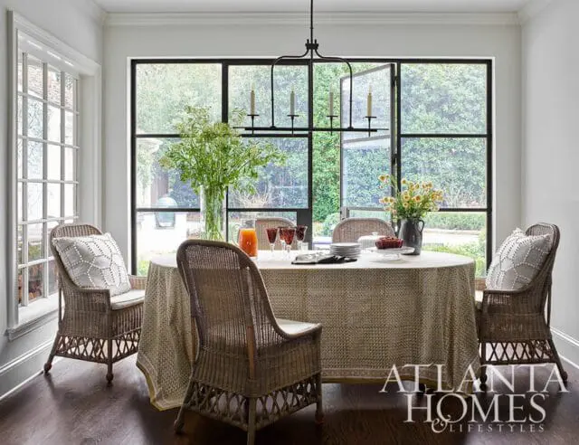Dining room with round table covered in patterned tablecloth, wicker chairs with patterned cushions, and large black-framed windows overlooking greenery.