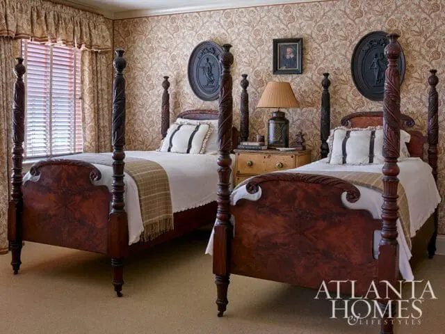 Bedroom with two ornate mahogany four-poster beds, patterned wallpaper, and floral curtains. A nightstand with a lamp sits between the beds.