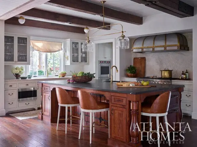 Kitchen with dark wood beams on ceiling, white cabinetry, and a large wooden island topped with a dark stone countertop. Leather bar stools with white bases surround the island.