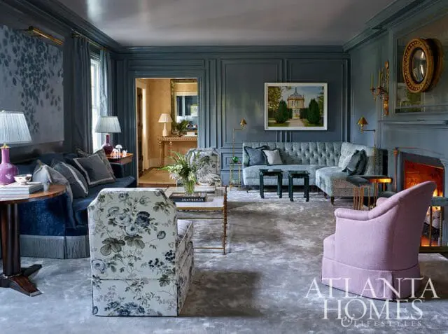 Blue living room with tufted gray sectional sofa, floral armchair, lavender accent chair, and paneled walls. A fireplace is visible on the right.