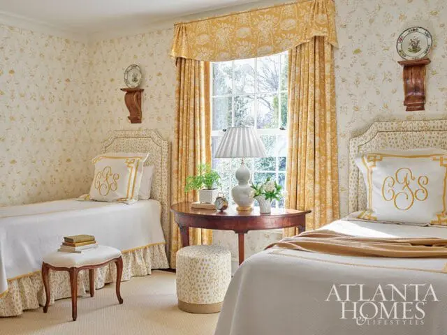 Bedroom with two twin beds, yellow floral wallpaper, yellow patterned curtains, and a round wooden side table with a lamp and flowers.